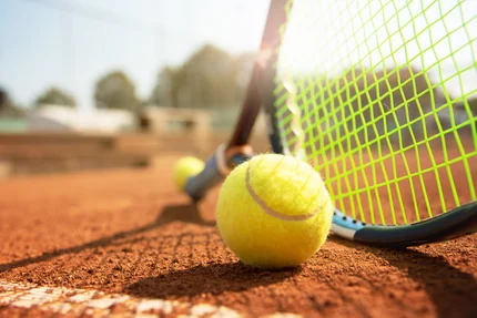 Ein Tennisball liegt auf einem Tennisplatz mit einem Schläger im Hintergrund. Der Schläger hat ein grünes Netz und die Sonne scheint. Der Platz ist aus rotem Lehm.