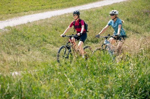 Zwei Frauen fahren auf Mountainbikes in einem grünen Feld. Ein Weg aus Kies führt hinter ihnen vorbei.