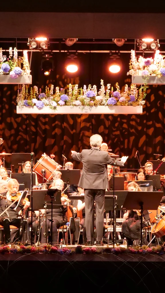 Ein Orchester spielt auf einer Bühne, während ein Dirigent die Musiker anleitet. Viele Instrumente sind sichtbar, und die Szene hat eine festliche Atmosphäre mit Blumenarrangements über der Bühne.