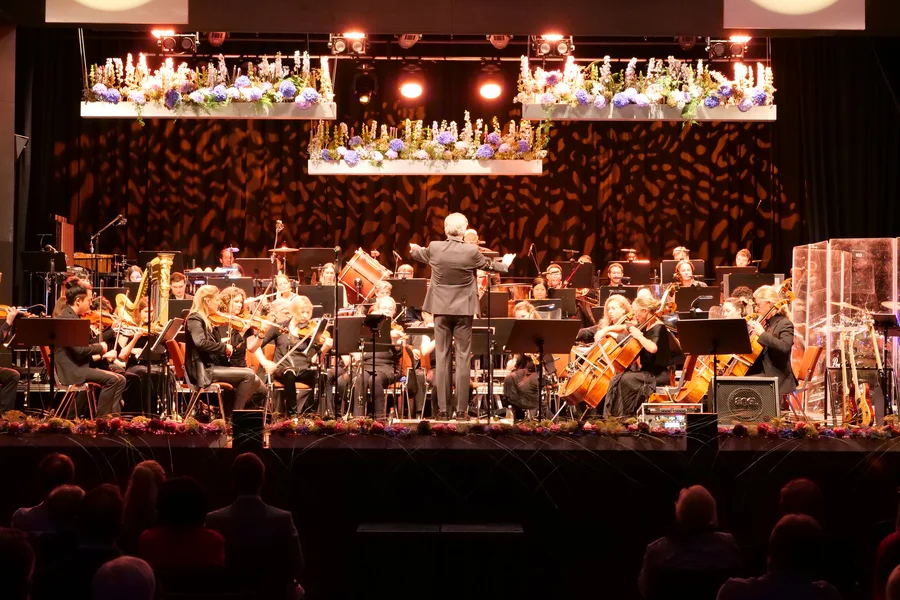 Orchesteraufführung mit Dirigent Ein Orchester spielt auf einer Bühne, während ein Dirigent die Musiker anleitet. Viele Instrumente sind sichtbar, und die Szene hat eine festliche Atmosphäre mit Blumenarrangements über der Bühne.