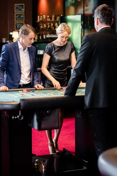 Zwei Personen konzentrieren sich auf einen Roulettetisch in einem Casino. Der Croupier erklärt die Regeln. Im Hintergrund steht ein weiterer Mann.