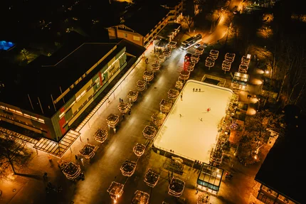 Stadt bei Nacht mit Eisbahn Nachtaufnahme einer beleuchteten Stadt mit Straßen, Gebäuden und einem großen Eisplatz, auf dem einige Menschen laufen. Bäume entlang der Wege sind mit Lichtern geschmückt.