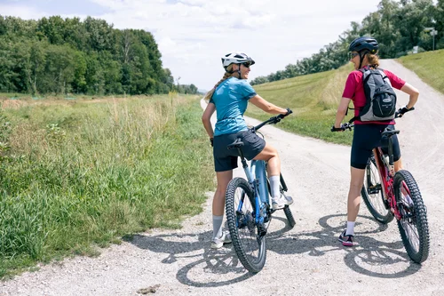 Zwei Radfahrerinnen stehen auf einem Waldweg. Die eine trägt ein blaues T-Shirt, die andere ein rotes. Sie unterhalten sich und blicken zurück. Umgeben von grasbewachsenen Flächen und Bäumen.