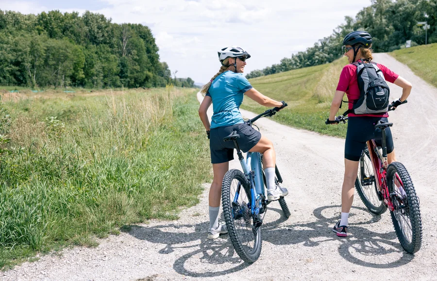 Zwei Radfahrerinnen stehen auf einem Waldweg. Die eine trägt ein blaues T-Shirt, die andere ein rotes. Sie unterhalten sich und blicken zurück. Umgeben von grasbewachsenen Flächen und Bäumen.