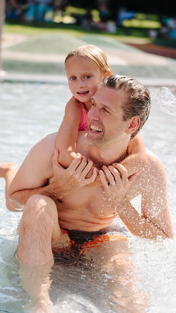 Ein Vater sitzt im Wasser mit seiner Tochter, die auf seinem Rücken sitzt. Beide lächeln und genießen das Spiel. Spritzendes Wasser ist im Hintergrund sichtbar. Es ist ein sonniger Tag.