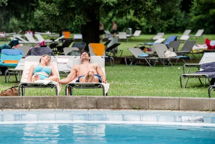 Ein Mann und eine Frau entspannen auf Liegestühlen am Pool umgeben von Sonnenliegen. Der Mann trägt eine Shorts, die Frau einen Badeanzug. Im Hintergrund sind weitere Sonnenliegen und ein grüner, bewaldeter Bereich sichtbar.