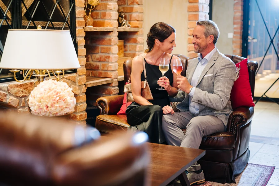 Ein elegant gekleidetes Paar sitzt auf einem Ledersofa und stößt mit zwei Gläsern Wein an.