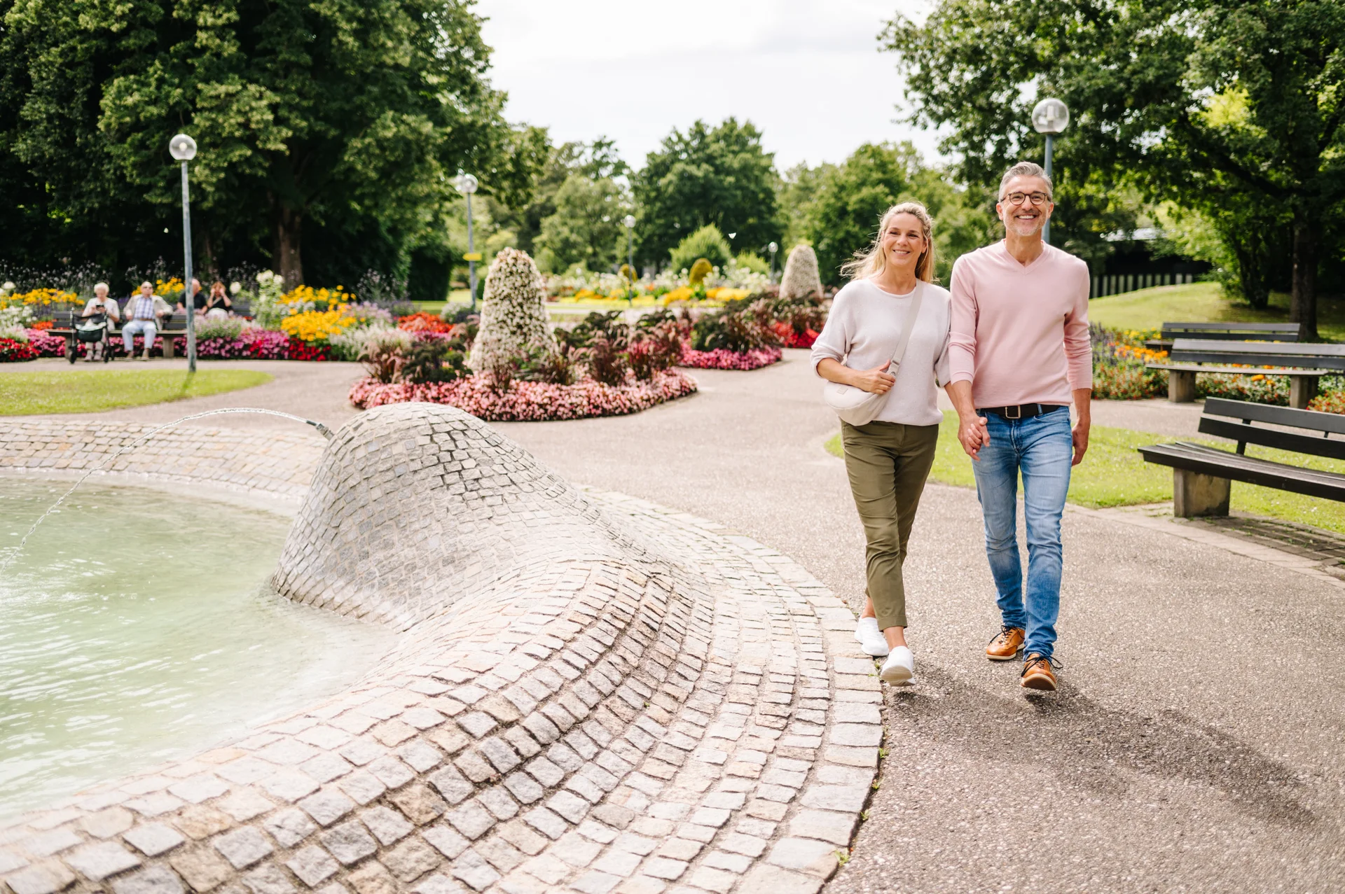 Ein Paar spaziert in einem Park mit bunten Blumen und einem Brunnen. Im Hintergrund sitzen Personen auf Bänken.