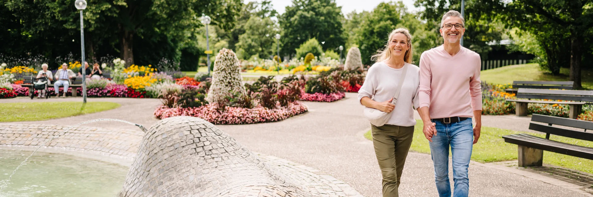 Ein Paar spaziert in einem Park mit bunten Blumen und einem Brunnen. Im Hintergrund sitzen Personen auf Bänken.