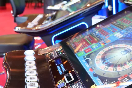 Spielautomaten im Casino Naheaufnahme von Spielautomaten in einem Casino mit bunten Lichtern und Bedienelementen. Im Hintergrund sind Tische und Stühle sichtbar.