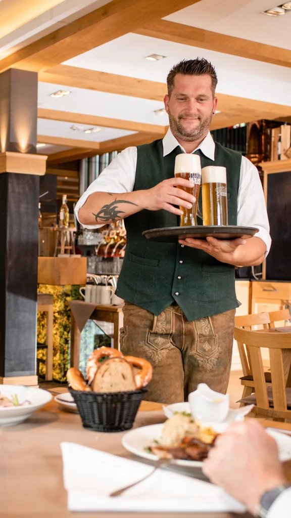 Kellner serviert Bier im Restaurant Ein Kellner serviert zwei Biergläser auf einem Tablett an Gäste in einem Restaurant. Eine Frau lächelt, während ein Mann am Tisch sitzt. Auf dem Tisch sind Essen und Blumen.