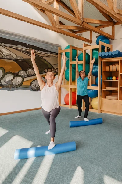 Vier Frauen absolvieren gemeinsam ein Fitnesstraining in einem Raum mit schrägen Holzdecken. Sie stehen auf blauen Schaumstoffrollen und haben die Arme in die Höhe gestreckt. Im Hintergrund sind sportliche Ausstattungen sichtbar.