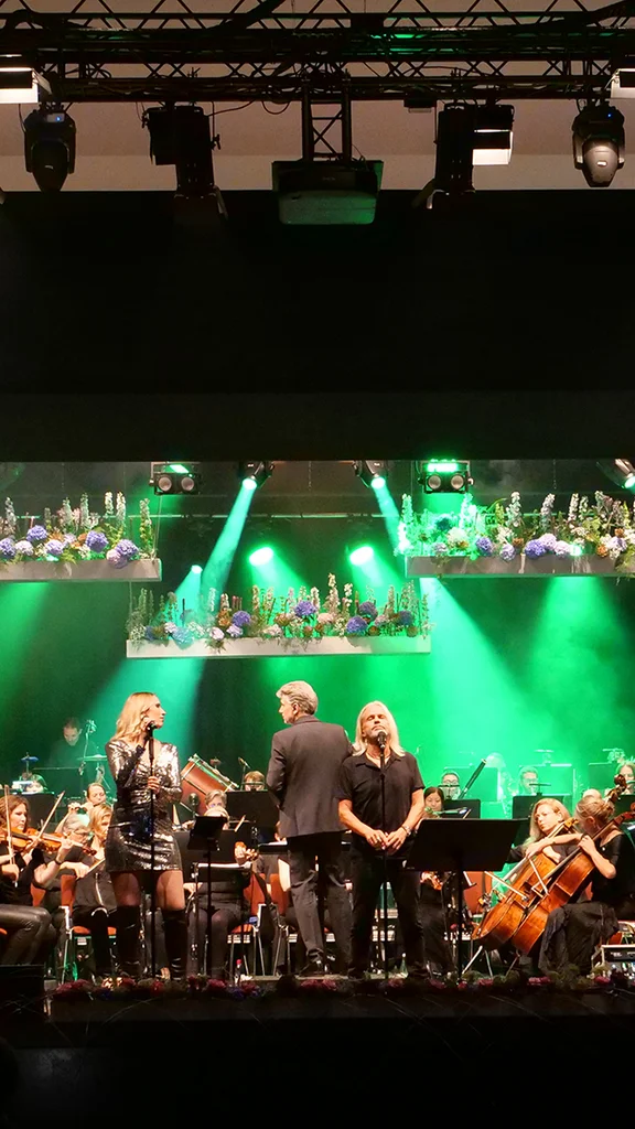 Eine Live-Aufführung mit Sängern und einem Orchester auf einer Bühne, mit grünen Lichtern und Blumenarrangements im Hintergrund.