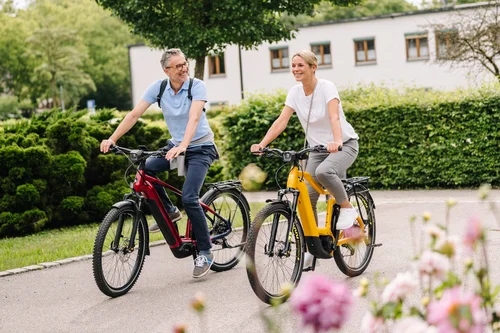 Ein Mann und eine Frau fahren auf Elektrofahrrädern in einem Park. Im Hintergrund sind Bäume und ein Gebäude sichtbar. Vorne blühen pinke Blumen.