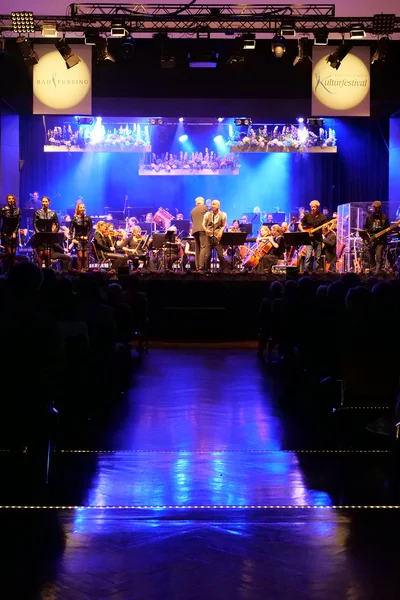 Orchestral Performance with Presenters A performance stage with musicians, two men speaking, and women in shiny outfits. The audience can be seen in the foreground. Colorful lights illuminate the stage.