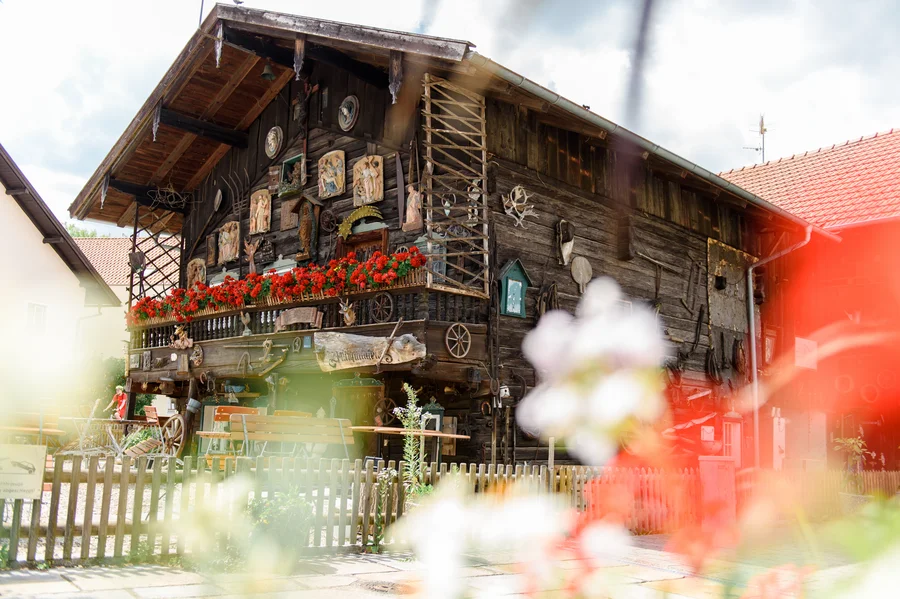 Traditionelles rustikales Haus Ein traditionelles, rustikales Haus mit Holzverkleidung, verzierten Fensterläden und Blumenkästen voller roter Geranien. Viele dekorative Gegenstände, darunter Figuren und Wandkunst, schmücken die Fassade. Ein gemütlicher Außenbereich mit Holztischen und -bänken ist sichtbar.