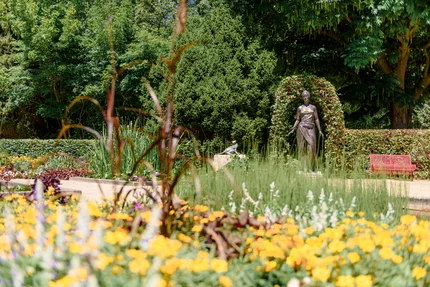 Ein Garten mit bunten Blumen und einer Statue, die von Bepflanzungen umgeben ist. Im Hintergrund sind grüne Büsche und ein roter Parkbank zu sehen.