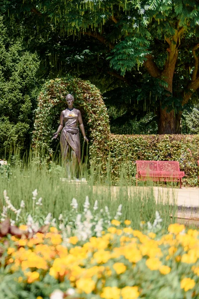 Ein Garten mit bunten Blumen und einer Statue, die von Bepflanzungen umgeben ist. Im Hintergrund sind grüne Büsche und ein roter Parkbank zu sehen.