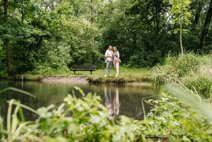 Zwei Frauen stehen am Ufer eines ruhigen Gewässers umgeben von Bäumen und Gras. Eine Bank ist im Hintergrund sichtbar.