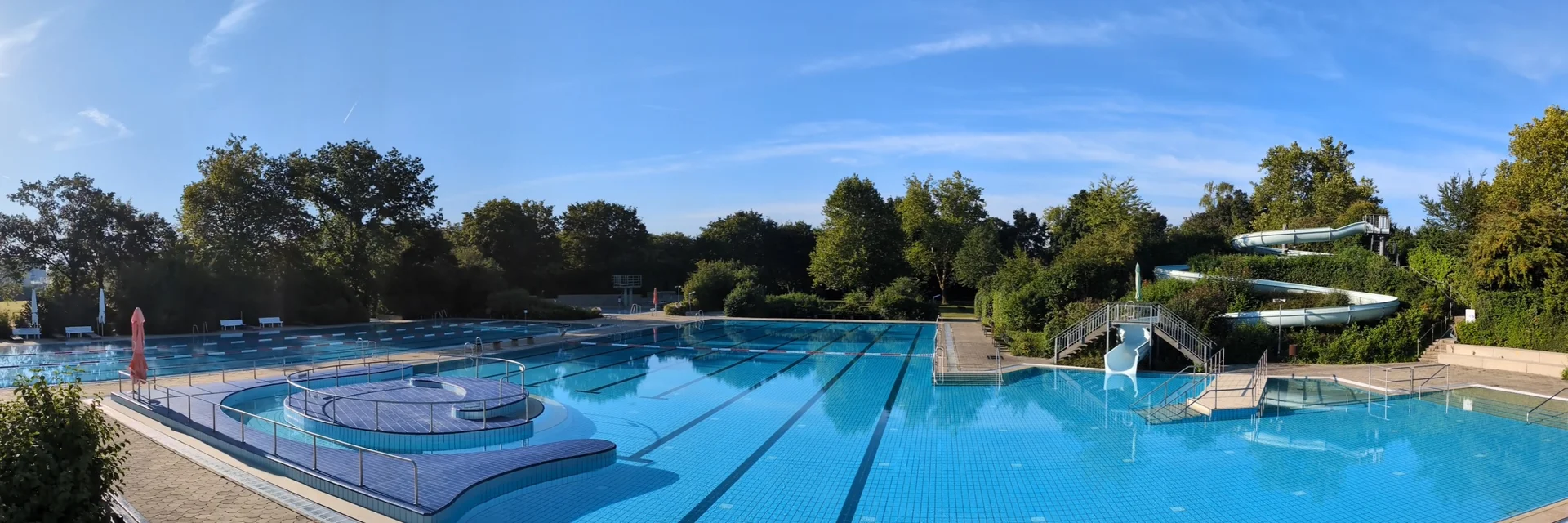 Ein Schwimmbad mit mehreren Becken, einer Rutsche und schattigen Bäumen im Hintergrund. Der Himmel ist klar und blau.