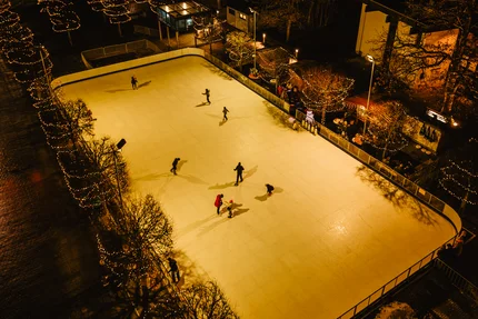 Beleuchtete Eisbahn bei Nacht Nachtaufnahme eines beleuchteten Eisstadions im Freien mit mehreren Schlittschuhläufern, umgeben von Bäumen mit Lichterketten und einigen Gebäuden.