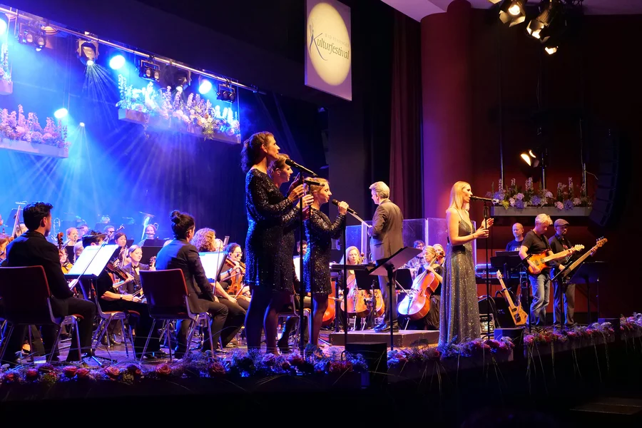 Ein spektakuläres Konzert mit Sängern und einem Orchester auf der Bühne, umgeben von bunten Lichtern und Blumenarrangements.