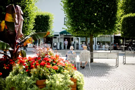 Ältere Menschen im Park Zwei ältere Menschen sitzen auf Stühlen in einem blühenden Park mit bunten Blumen und Bäumen. Im Hintergrund ist ein Cafe zu sehen.