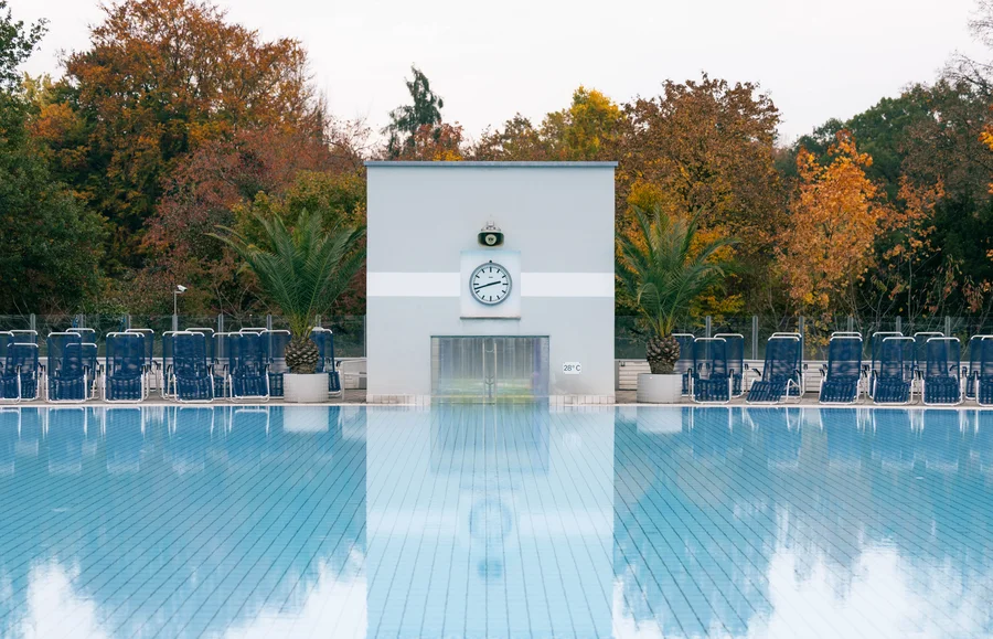Pool Reflection with Clock Ein Pool mit blauen Fliesen spiegelt die Umgebung wider. Dahinter steht ein graues Gebäude mit einer Uhr und einigen Palmenn. Gestapelte Liegestühle sind an einer Seite des Pools angeordnet.