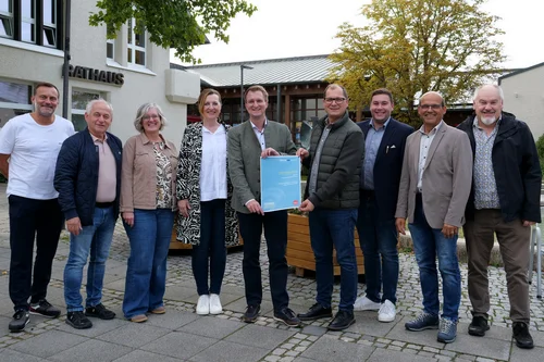 Gruppe vor dem Rathaus Neun Personen stehen vor einem Rathaus und halten gemeinsam ein blaues Dokument in die Kamera.