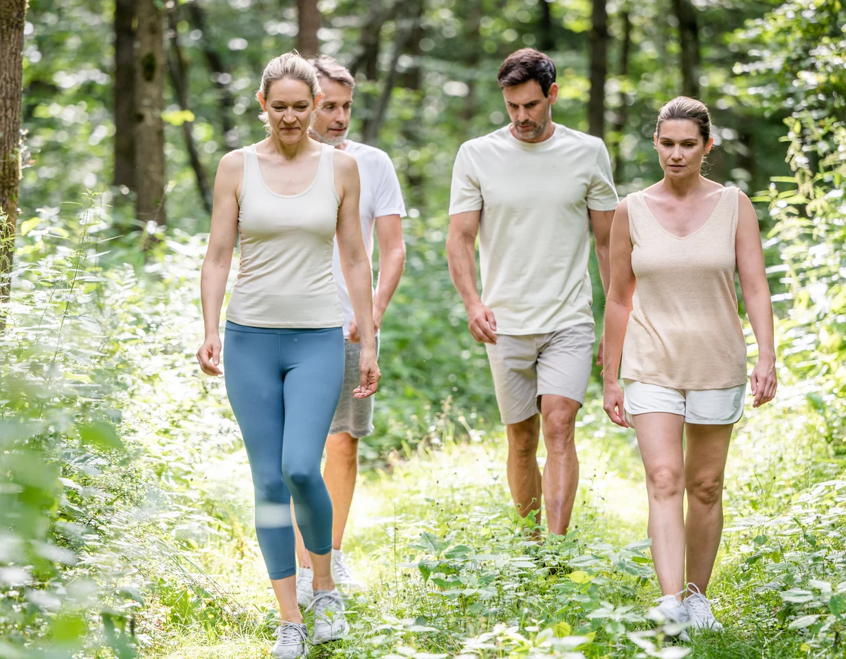 Eine Gruppe von vier Personen geht auf einem Waldweg. Sie tragen legere Sportkleidung und scheinen sich in der Natur aufzuhalten.