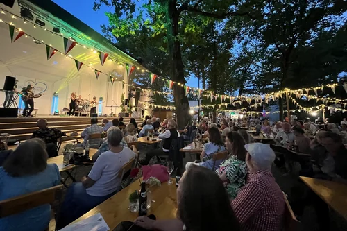 Evening Outdoor Concert Performance A lively outdoor performance takes place in the evening, with a band on stage and an audience seated at wooden tables. Colorful bunting and string lights illuminate the scene, creating a festive atmosphere.