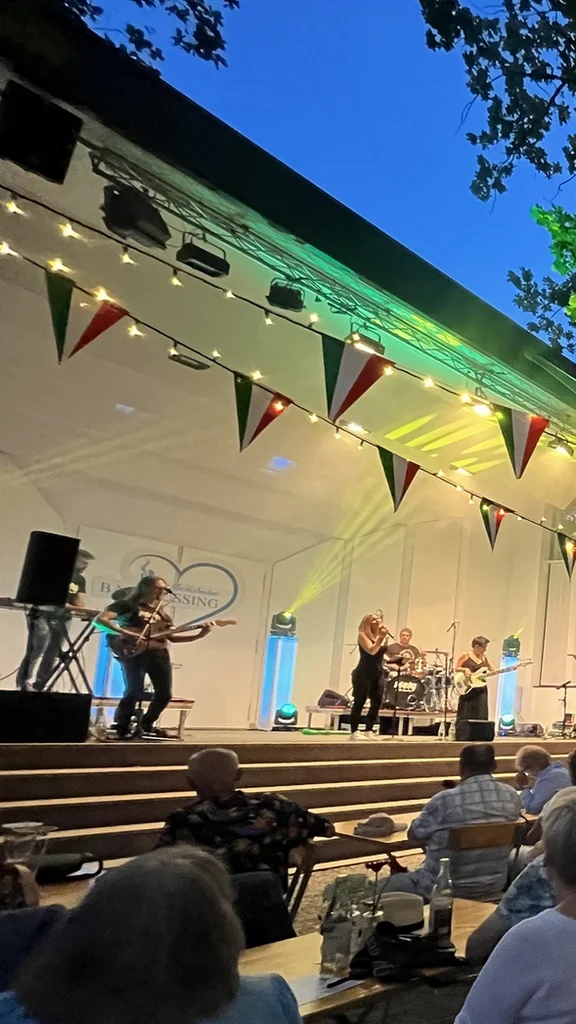 A lively outdoor performance takes place in the evening, with a band on stage and an audience seated at wooden tables. Colorful bunting and string lights illuminate the scene, creating a festive atmosphere.