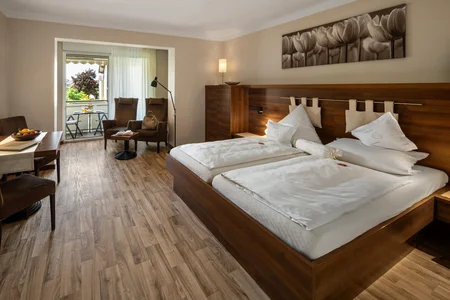 Ein helles Zimmer mit zwei Einzelbetten, einem Tisch und Stühlen. Im Hintergrund ist ein Balkon mit einem Tisch und zwei Stühlen, der auf einen grünen Bereich blickt. Es gibt eine Lampe und moderne Möbel.