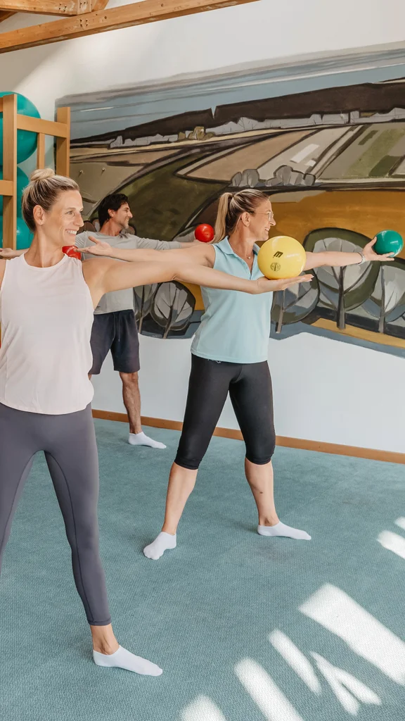 Gruppentraining mit Fitnessbällen Fünf Personen führen in einem Fitnessraum Armübungen mit Fitnessbällen aus, während sie nebeneinander stehen. Im Hintergrund sind Fitnessgeräte und eine Wandmalerei zu sehen.