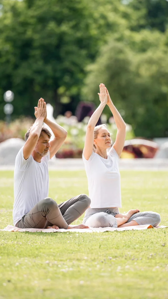 Drei Menschen sitzen im Gras auf Yogamatten, meditieren mit erhobenen Händen. Es ist ein sonniger Tag im Park mit blühenden Pflanzen im Hintergrund.