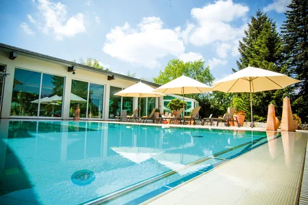 Ein Poolbereich mit Liegestühlen und Sonnenschirmen, umgeben von Pflanzen und einem modernen Gebäude im Hintergrund unter blauem Himmel.