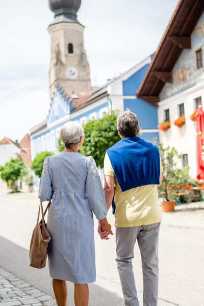 Ein älteres Paar spaziert Hand in Hand auf einer ruhigen Straße mit bunten Gebäuden und Bäumen. Im Hintergrund ist ein Kirchturm sichtbar.