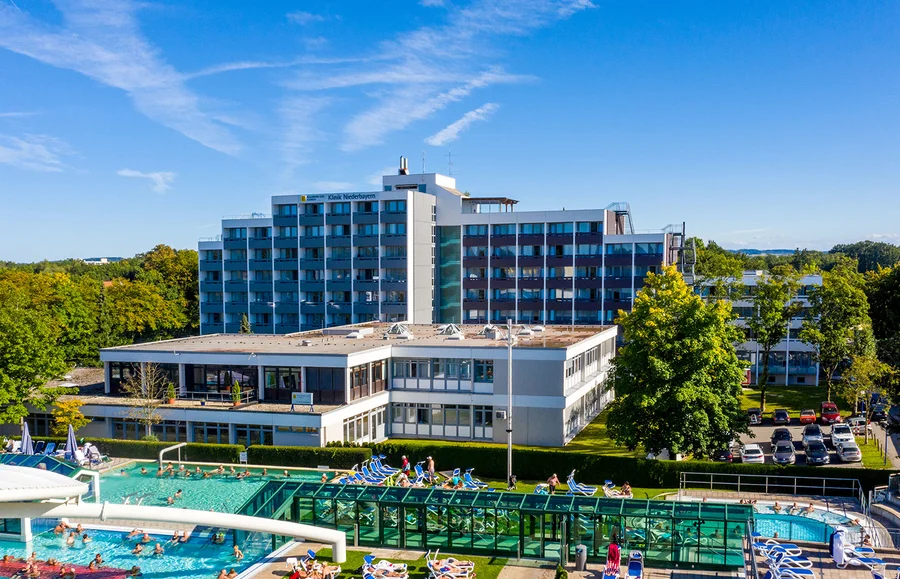 Ein Blick auf die Klinik Niederbayern mit einem Poolbereich und Liegen davor. Viele Menschen genießen den sonnigen Tag am Pool und auf dem Rasen. Umgeben von Bäumen und einem Parkplatz.