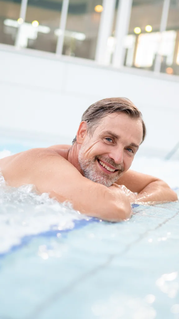 Ein lächelnder Mann liegt im Wasser eines Schwimmbeckens, mit den Armen auf dem Beckenrand. Er hat graue Haare und trägt kein Oberteil. Im Hintergrund sind große Fenster und sanierte Wände zu sehen.