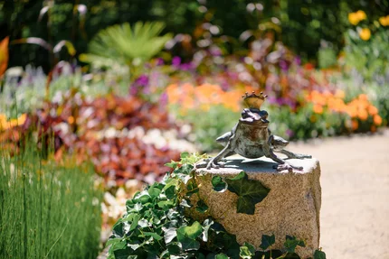 Eine Statue eines Frosches mit einer Krone, die auf einem Stein sitzt, umgeben von bunten Blumen und Pflanzen im Garten.