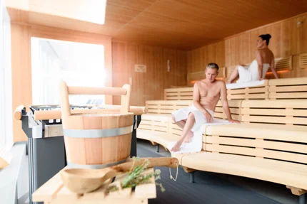 In a sauna, a wooden bucket and scoop are in the foreground. A shirtless man sits on a wooden bench, while a woman in a white towel relaxes in the background.