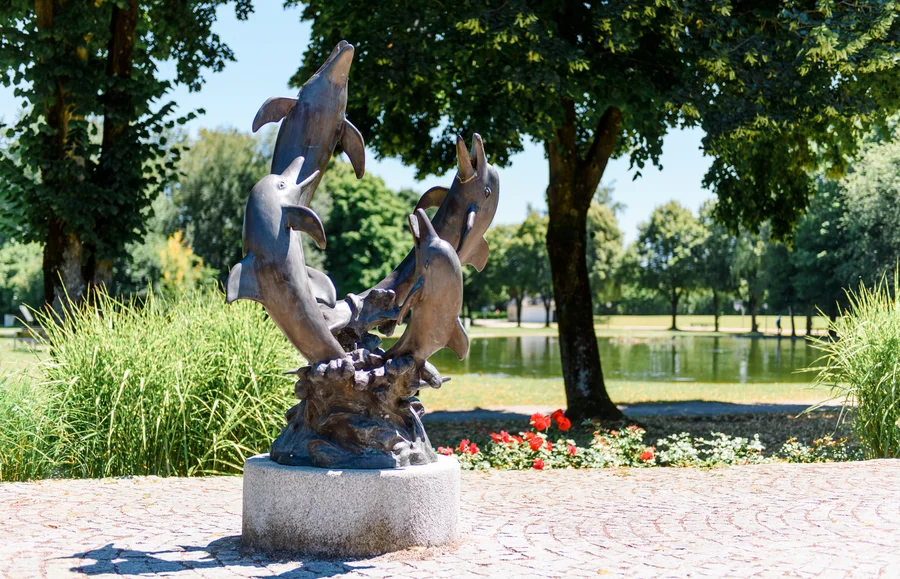 Eine Bronzeskulptur von Delfinen, die in einer dynamischen Pose angeordnet sind. Sie springenden aus dem Wasser, umgeben von grünen Pflanzen und Bäumen, im Hintergrund ein ruhiges Gewässer.