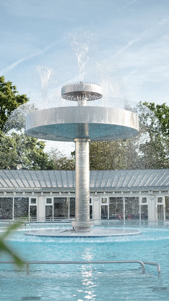 Ein moderner Springbrunnen in einem Schwimmbecken, umgeben von Bäumen und einem Gebäude mit Glasfenstern. Wasser sprüht spielerisch von der obersten Plattform des Brunnens.