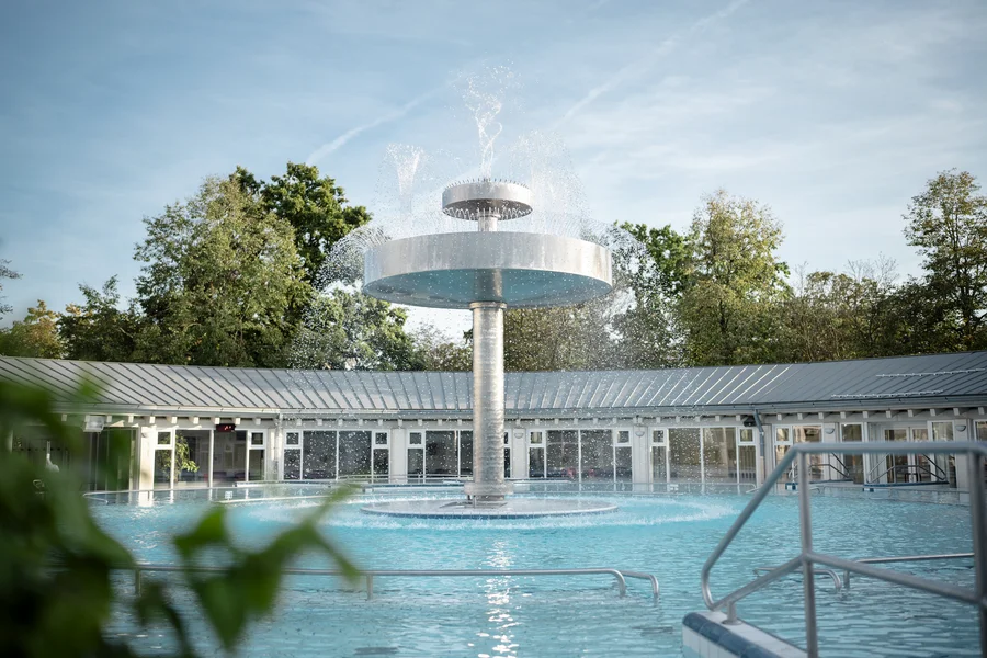 Ein moderner Springbrunnen in einem Schwimmbecken, umgeben von Bäumen und einem Gebäude mit Glasfenstern. Wasser sprüht spielerisch von der obersten Plattform des Brunnens.