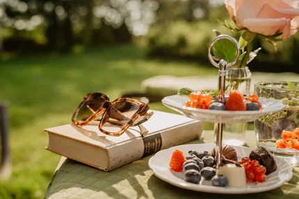 Ein Tischgedeck im Freien mit einem Buch, Sonnenbrille, zwei Etagere mit Früchten und Schokolade sowie einer Blume in einer Vase.