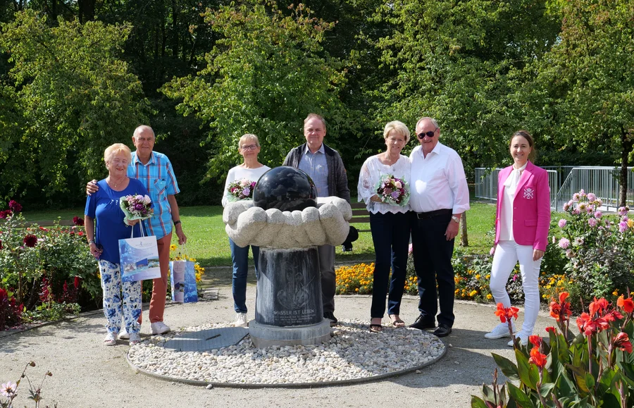 Sechs Erwachsene stehen in einem Garten um einen Brunnen mit schwarzer Kugel; mehrere halten Blumensträuße und Geschenktüten. Im Hintergrund sind Bäume, Blumenbeete und ein Geländer zu sehen.