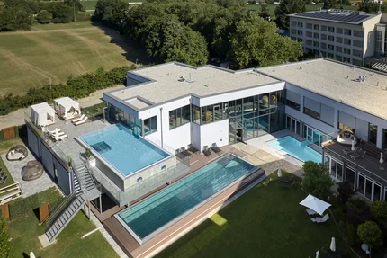 Moderne Villa mit Pools Eine moderne Villa mit zwei Pools, Liegen und Sonnenschirmen umgeben von Rasenflächen und Bäumen. Ein geschützter Terrassebereich ist sichtbar, und im Hintergrund steht ein Wohngebäude.
