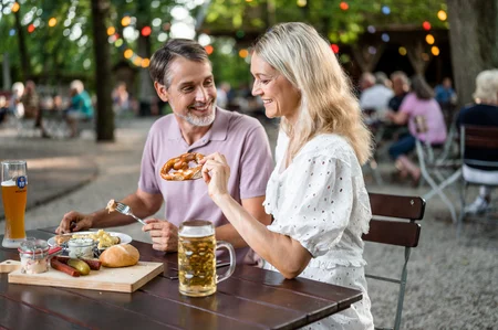 Gemütliches Essen im Freien Ein Mann und eine Frau genießen ein Essen im Freien. Der Mann hält eine Gabel mit Fleisch, während die Frau ein Brezel in der Hand hat. Auf dem Tisch sind weitere Speisen und Getränke.