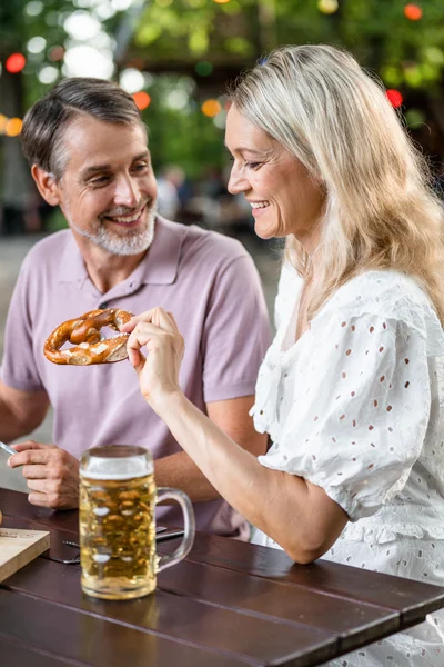 Ein Mann und eine Frau genießen ein Essen im Freien. Der Mann hält eine Gabel mit Fleisch, während die Frau ein Brezel in der Hand hat. Auf dem Tisch sind weitere Speisen und Getränke.