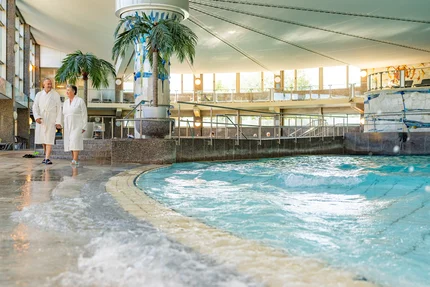 Innenpool mit Paar Ein älteres Paar im Bademantel steht in einer Halle neben einem Innenpool mit sprudelndem Wasser und Palmen. Im Hintergrund sind große Fenster, Geländer und weitere Gäste zu sehen.
